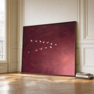 May include: A large rectangular wall art piece featuring a flock of white birds flying in a V-formation against a deep, solid burgundy red background. The painting is framed in a thin dark brown border and rests on a wooden floor.