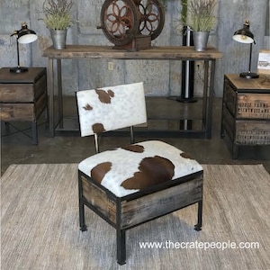 K&ouml;nnte beinhalten: Ein rustikaler Holz-Kistenhocker mit einer braunen und wei&szlig;en Kuhfell-Sitzfl&auml;che. Der Hocker wird von The Crate People hergestellt und hat einen Metallrahmen. Der Hocker steht auf einem Teppich vor einem Holztisch mit einer antiken Kaffeem&uuml;hle darauf.