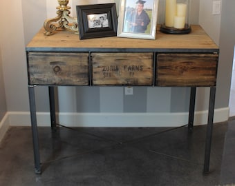 Puede incluir: Una mesa consola de madera rústica con patas de metal y tres cajones. Los cajones están etiquetados con "Zoria Farms, San Francisco, CA". La mesa está decorada con un candelabro dorado, un arreglo de flores blancas, dos fotos enmarcadas y una campana de cristal con velas.