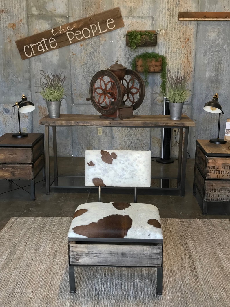 K&ouml;nnte beinhalten: Ein rustikaler Holzstuhl mit einer Kuhfell-Sitzfl&auml;che und -R&uuml;ckenlehne. Der Stuhl steht auf einem gewebten Teppich vor einer Metallwand mit einem Schild, auf dem "the crate people" steht.
