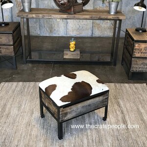 K&ouml;nnte beinhalten: Ein brauner und wei&szlig;er Kuhfell-Ottomane mit einem Holzrahmen und schwarzen Metallbeinen. Der Ottomane steht auf einem hellgrauen Teppich. Der Text "www.thecratepeople.com" ist in der unteren rechten Ecke des Bildes sichtbar.