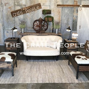 K&ouml;nnte beinhalten: Ein wei&szlig;es, gekn&ouml;pftes Sofa mit zwei Kuhfellst&uuml;hlen mit Holz- und Metallrahmen. Die St&uuml;hle haben Stauf&auml;cher. Die St&uuml;hle und das Sofa stehen vor einer rustikalen Wand mit einem Schild, auf dem "The Crate People" und eine Website-Adresse stehen.