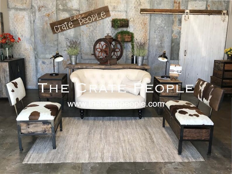 K&ouml;nnte beinhalten: Ein wei&szlig;es, gekn&ouml;pftes Sofa mit zwei Kuhfellst&uuml;hlen mit Holz- und Metallrahmen. Die St&uuml;hle haben Stauf&auml;cher. Die St&uuml;hle und das Sofa stehen vor einer rustikalen Wand mit einem Schild, auf dem "The Crate People" und eine Website-Adresse stehen.