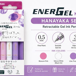 以下が含まれることがあります： 0.5mmの極細ペン先を持つ、ぺんてるエナージェル・コハレ・ハナヤカセットのゲルインキボールペン3本セット。パッケージには、ブロッサムピンク、サクラホワイト、ライラックパープルのペンが並んでおり、ホワイトのペンには黒インク、他のペンにはカラーインクが使用されています。