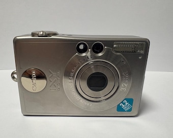 May include: A silver Canon IXY Digital compact camera featuring a 3.2 megapixel lens with 5.4-8.3mm focal length and f/2.8-4.0 aperture. The front includes a flash, a circular lens housing, and a blue sticker indicating 3.2 megapixels.