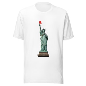 Pode incluir: Camiseta branca com uma imagem da Estátua da Liberdade segurando um cartão vermelho. A estátua é verde, em pé sobre uma base marrom. O cartão vermelho é levantado, criando um contraste visual marcante.