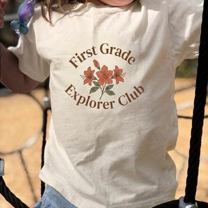 May include: A cream-colored short-sleeve t-shirt featuring a brown circular graphic with the text "First Grade Explorer Club" surrounding a central illustration of three orange flowers with green leaves and stems centered on the chest.