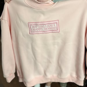 May include: A light pink sweatshirt with a crew neck and long sleeves. The front features an embroidered rectangular patch with the text "HOT GIRLS HAVE GRANNY HOBBIES" in a pink and white design. The sweatshirt is made of a soft material.