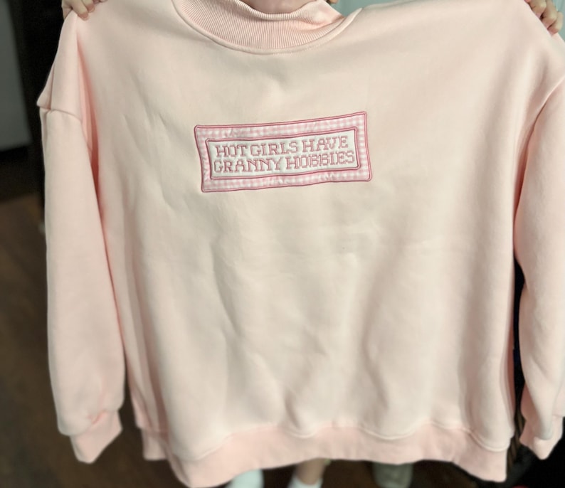 May include: A light pink sweatshirt with a crew neck and long sleeves. The front features an embroidered rectangular patch with the text "HOT GIRLS HAVE GRANNY HOBBIES" in a pink and white design. The sweatshirt is made of a soft material.