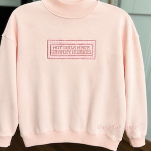 May include: Pale pink sweatshirt with a high neck. It has an embroidered rectangular patch on the front, with the text "HOT GIRLS HAVE GRANNY HOBBIES" in a serif font. The patch is framed by a gingham border.