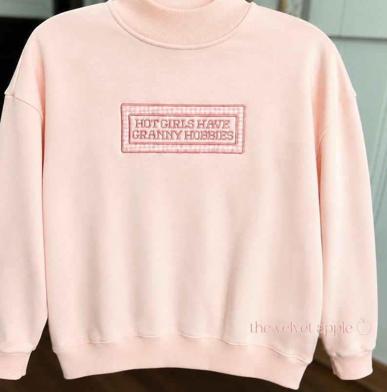 May include: A light pink sweatshirt with a mock neck. The front features an embroidered rectangular patch with the text "HOT GIRLS HAVE GRANNY HOBBIES" in a serif font. The patch has a gingham border.