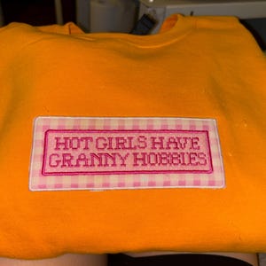 May include: An orange sweatshirt with a rectangular patch that reads "HOT GIRLS HAVE GRANNY HOBBIES" in pink embroidery. The patch has a pink and white gingham border.
