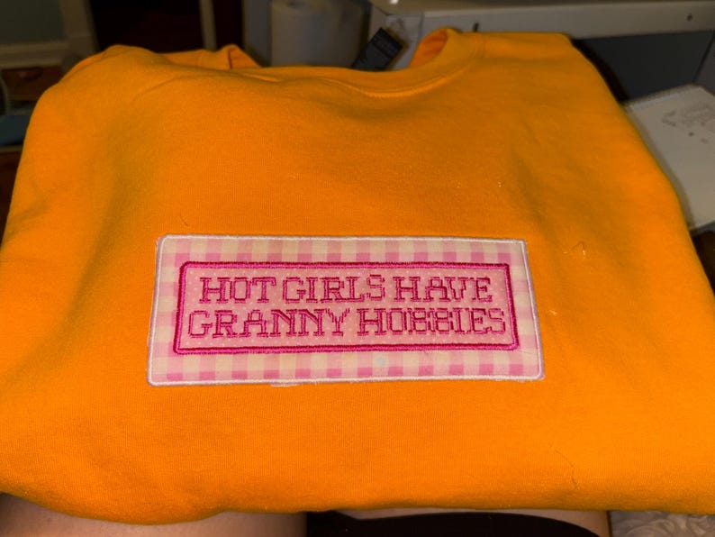 May include: An orange sweatshirt with a rectangular patch that reads "HOT GIRLS HAVE GRANNY HOBBIES" in pink embroidery. The patch has a pink and white gingham border.