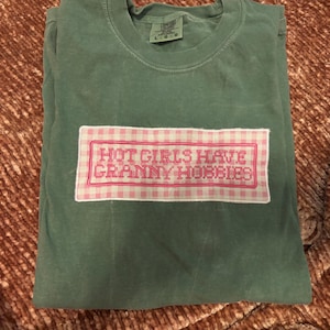 May include: A folded, sage green t-shirt with a rectangular patch featuring the text "HOT GIRLS HAVE GRANNY HOBBIES" in pink embroidery on a pink and white gingham background. The shirt is made of a soft material.