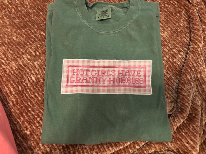 May include: A folded, sage green t-shirt with a rectangular patch featuring the text "HOT GIRLS HAVE GRANNY HOBBIES" in pink embroidery on a pink and white gingham background. The shirt is made of a soft material.