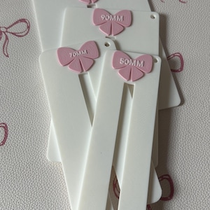 May include: A set of white rectangular plastic templates with a vertical slot, each featuring a pink bow accent at the top. The bows are embossed with the text 50MM, 70MM, 90MM, and 110MM, indicating the specific size of each individual tool.