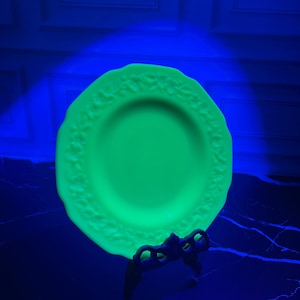 May include: A round, bright neon green glass plate featuring a decorative embossed floral pattern along its scalloped rim. The plate is displayed upright on a black metal stand, glowing intensely under ultraviolet light in a dark environment.