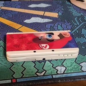 May include: A white handheld gaming console featuring a graphic of Mario on the top cover. The device is closed and displays a red and blue color scheme on the lid with the character's face and signature red hat visible on the right side.