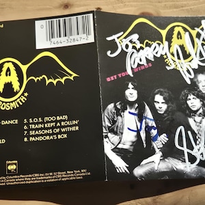 Puede incluir: Portada del álbum en CD "Get Your Wings" de Aerosmith. La portada del álbum es negra con el logotipo de la banda en amarillo. La caja del CD está abierta, revelando las firmas de los miembros de la banda y una foto de la banda.