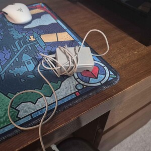 May include: A light gray rectangular power adapter with a coiled white cord. The adapter is positioned on a desk mat featuring a colorful stained-glass style pattern of a castle and a heart, alongside a white computer mouse with a green logo.