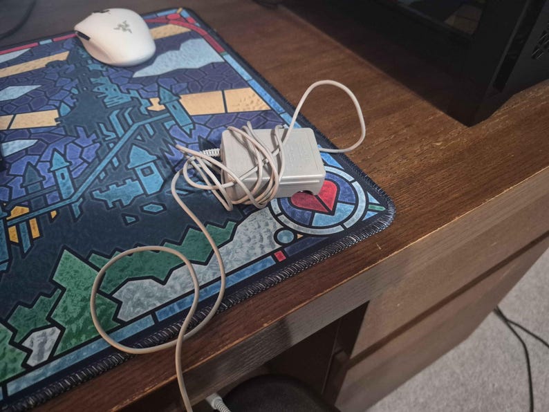 May include: A light gray rectangular power adapter with a coiled white cord. The adapter is positioned on a desk mat featuring a colorful stained-glass style pattern of a castle and a heart, alongside a white computer mouse with a green logo.
