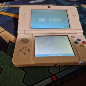 May include: A white handheld gaming console with a dual-screen display, a directional pad, a circular analog stick, and four colored action buttons labeled X, Y, A, and B. The bottom screen displays the text Create Folder with two glowing indicator lights.