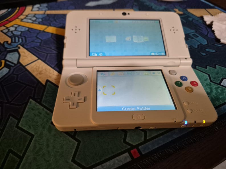 May include: A white handheld gaming console with a dual-screen display, a directional pad, a circular analog stick, and four colored action buttons labeled X, Y, A, and B. The bottom screen displays the text Create Folder with two glowing indicator lights.