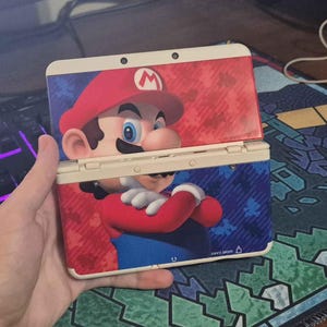 May include: A handheld gaming console featuring a graphic of Mario on the exterior shell. The device has a cream-colored frame and a red and blue background pattern with small Mario silhouettes. The console is shown in an open, upright position.