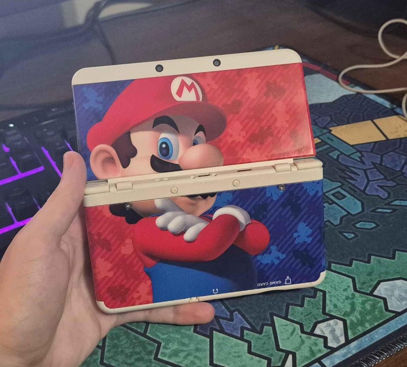 May include: A handheld gaming console featuring a graphic of Mario on the exterior shell. The device has a cream-colored frame and a red and blue background pattern with small Mario silhouettes. The console is shown in an open, upright position.