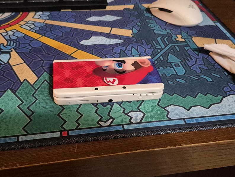 May include: A white handheld gaming console featuring a graphic of Mario on the top cover. The device is closed and displays a red and blue color scheme on the lid with the character's face and signature red hat visible on the right side.