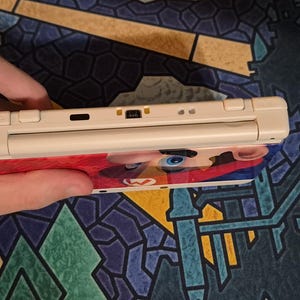 May include: A handheld gaming console featuring a cream-colored top edge with shoulder buttons, a charging port, and an infrared sensor. The device displays a vibrant graphic of a character with a red hat and blue eyes on its outer shell.