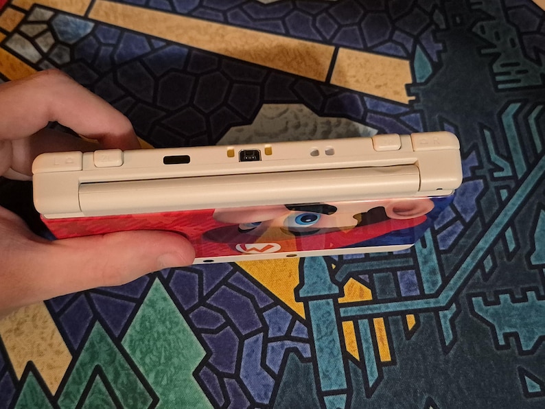 May include: A handheld gaming console featuring a cream-colored top edge with shoulder buttons, a charging port, and an infrared sensor. The device displays a vibrant graphic of a character with a red hat and blue eyes on its outer shell.