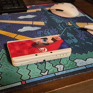 May include: A white handheld gaming console featuring a graphic of Mario on the top cover. The device is closed and displays a red and blue color scheme with a prominent image of Mario's face and hat. The console has a rectangular shape with visible ports.
