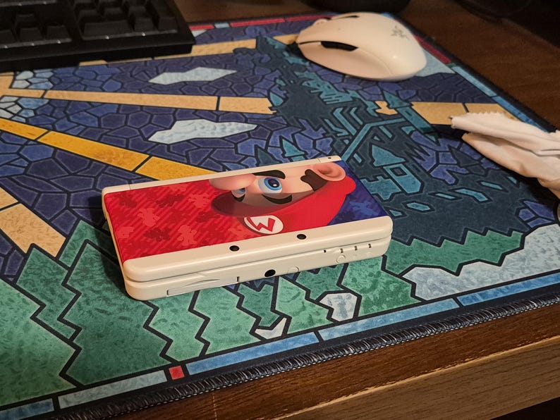May include: A white handheld gaming console featuring a graphic of Mario on the top cover. The device is closed and displays a red and blue color scheme with a prominent image of Mario's face and hat. The console has a rectangular shape with visible ports.