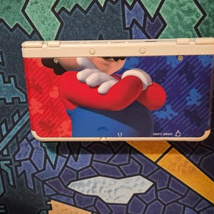 May include: A white handheld gaming console featuring a graphic of Mario with crossed arms on the front cover. The design includes a red and blue background with small silhouettes of the character. The text GAME CARD is printed near the bottom right.