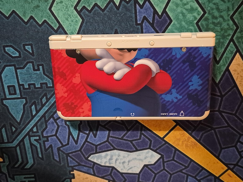 May include: A white handheld gaming console featuring a graphic of Mario with crossed arms on the front cover. The design includes a red and blue background with small silhouettes of the character. The text GAME CARD is printed near the bottom right.
