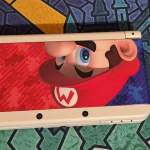 May include: A white handheld gaming console featuring a graphic of Mario on the top cover. The design includes a red background with small silhouettes of Mario, a large close-up of Mario's face wearing a red cap with a white W, and a blue section.