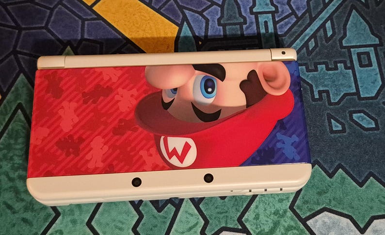 May include: A white handheld gaming console featuring a graphic of Mario on the top cover. The design includes a red background with small silhouettes of Mario, a large close-up of Mario's face wearing a red cap with a white W, and a blue section.