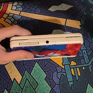 May include: A white handheld gaming console featuring a game card slot, a headphone jack, and a power indicator light on the top edge. The device has a colorful graphic skin applied to the front, displaying a red and blue character design.