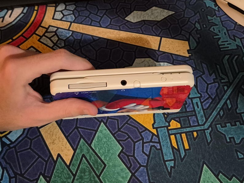 May include: A white handheld gaming console featuring a game card slot, a headphone jack, and a power indicator light on the top edge. The device has a colorful graphic skin applied to the front, displaying a red and blue character design.