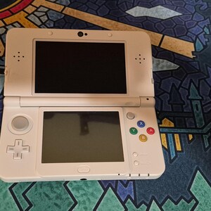 May include: A white handheld gaming console featuring a dual-screen design, a directional pad, a circular analog stick, and four colored action buttons labeled A, B, X, and Y. The device includes a central touch screen and a top display screen.