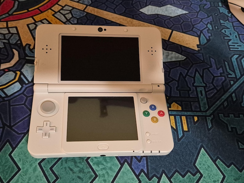 May include: A white handheld gaming console featuring a dual-screen design, a directional pad, a circular analog stick, and four colored action buttons labeled A, B, X, and Y. The device includes a central touch screen and a top display screen.