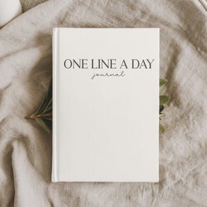 May include: A cream-colored journal with the text "ONE LINE A DAY journal" printed in black. The journal is lying on a neutral-toned fabric, with a sprig of greenery in the corner.