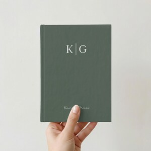 May include: A hand holding a muted green hardcover journal. The cover displays the initials "K | G" in white, with the script text "King Grace" below. The journal has a minimalist design and a sophisticated aesthetic.
