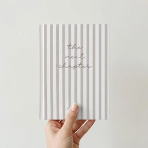 May include: A hardcover journal with a striped pattern in gray and white. The words "the next chapter" are written in cursive on the cover. The journal is held in a person's hand.