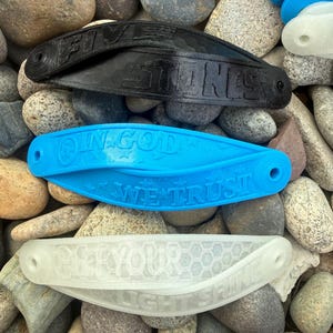 May include: Three curved, elongated plastic bands with holes at each end. The top band is black with the text "FIVE STONES". The middle band is bright blue with the text "IN GOD WE TRUST". The bottom band is translucent white with "LET YOUR LIGHT SHINE".