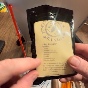May include: A black pouch containing a Sling Along Kit with a beige label. The label features the text Five Stones Slings, an inventory list, and the Bible verse Joshua 1:9. The kit includes a pouch, release cord set, retention cord set, button, cracker, release armor, and release beads.