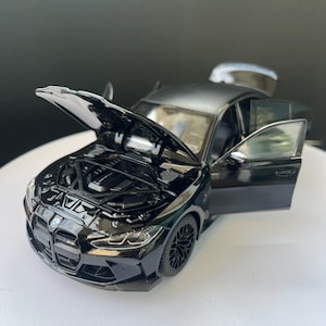 May include: A black die-cast model car with the bonnet and doors open, revealing engine details and interior. The car features black rims and a BMW emblem. The model is displayed on a white surface.