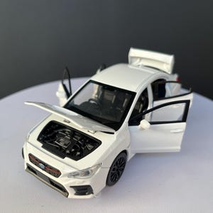 May include: A white die-cast model car featuring an open bonnet, open front doors, and an open boot. The vehicle has black wheels, a black front grille with a red accent line, and a visible engine bay. The interior is black with detailed seating.