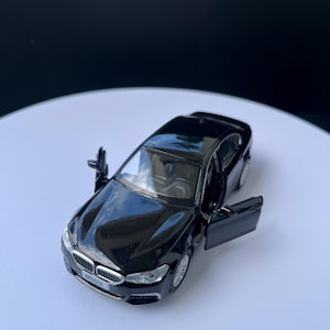 Könnte beinhalten: Ein schwarzes Modellauto mit geöffneten Türen, das den Innenraum zeigt. Das Auto hat silberne Felgen und einen Frontgrill mit dem Text "M550i". Das Auto steht auf einer weißen Oberfläche.